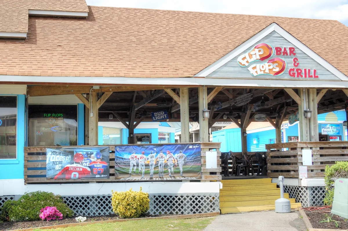 This image showcases the lively exterior of 'Flip Flops Bar & Grill,' with colorful banners, a prominent sign, and plenty of wooden deck seating. The scene is set up for people dining outdoors and socializing, suggesting a fun, communal vibe perfect for group activities. TVs for watching sports are visible, and the open-air setup hints at beach proximity and relaxed evenings out.