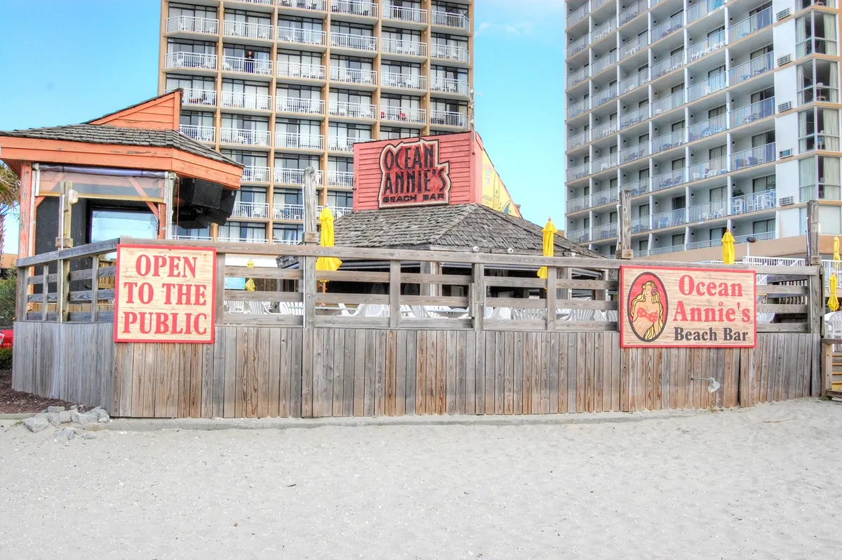 An inviting beachfront bar with signs for Ocean Annie’s and an 'open to the public' message sits against a backdrop of high-rise resorts and the beach. The open structure suggests a festive yet relaxed atmosphere ideal for socializing, live music, and beachfront cocktails.