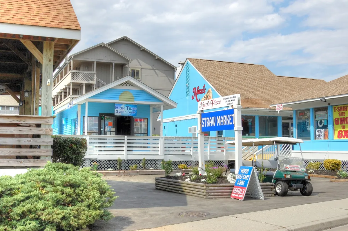 A lively, colorful scene at the Straw Market with signs for Nina's Pizza, High Tide Smoke Shop, and other local businesses. Families are seen strolling, dining outdoors, and discovering unique shops in a casual, welcoming environment.