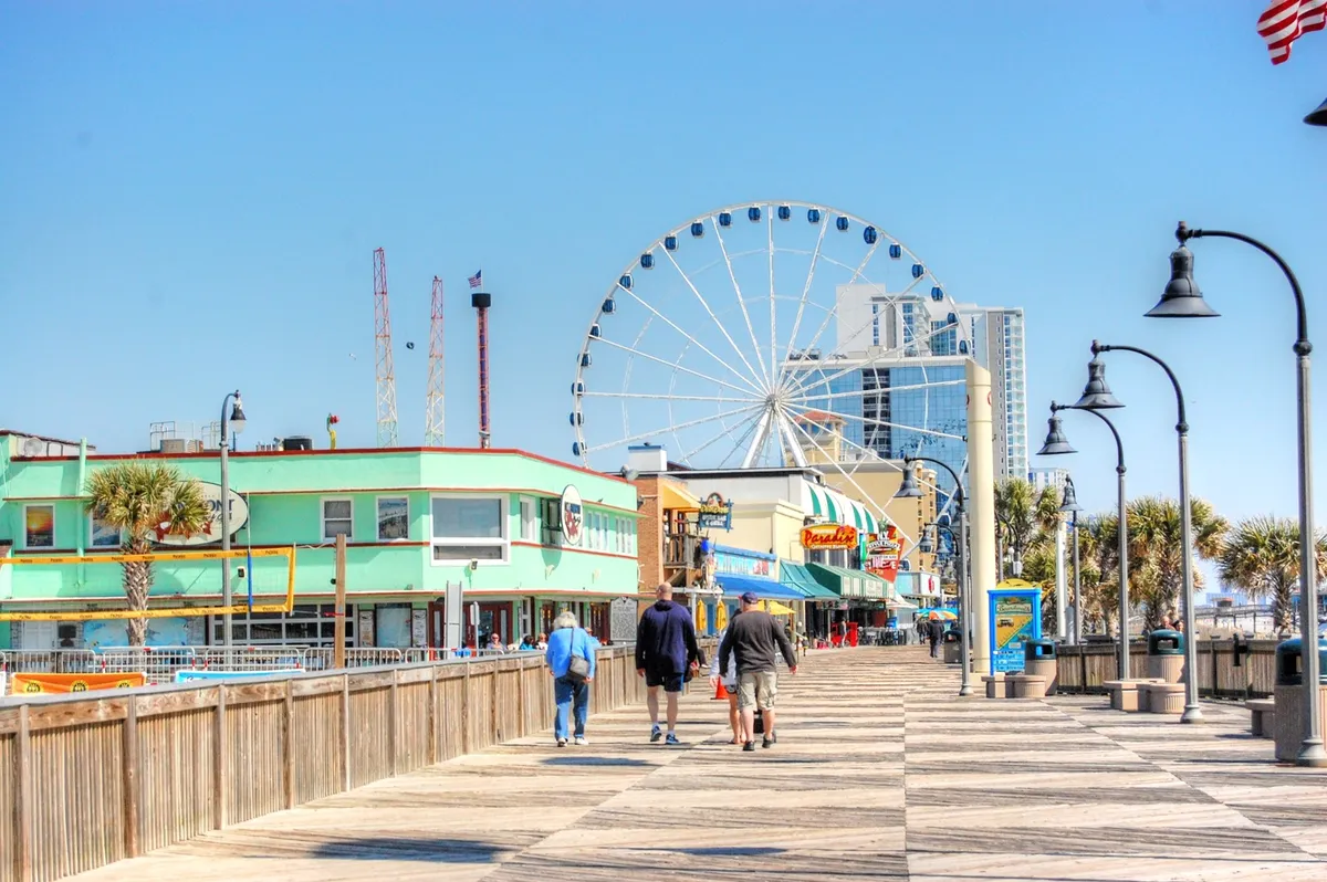 A lively boardwalk scene featuring a prominent ferris wheel, colorful storefronts, and people leisurely strolling. The image exudes a joyful and relaxed atmosphere, highlighting Myrtle Beach’s vibrant local attractions and the opportunity to enjoy amusement rides, shopping, and local treats.