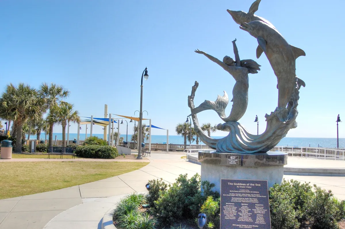 This scenic exterior image displays the iconic 'Goddess of the Sea' sculpture framed by palm trees, with the ocean visible in the background. It conveys a joyful, memorable moment of friends or groups strolling along the Myrtle Beach promenade, enjoying seaside art and natural beauty.