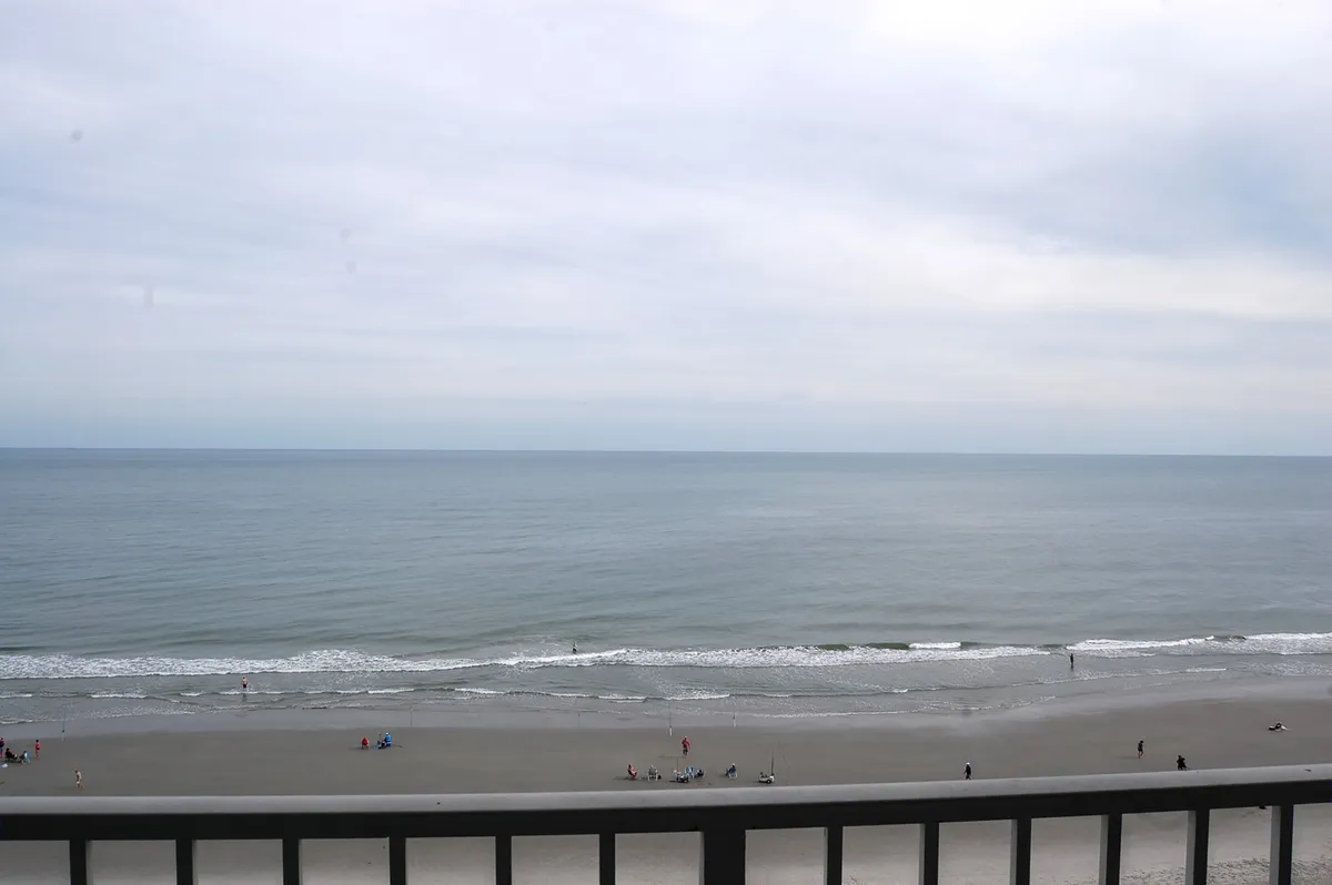 A wide-angle view of the beach shows the gentle stretch of sand, calm ocean waters, and a distant horizon. People can be seen strolling along the shore and enjoying beach activities, adding a subtle sense of exploration and possibility.