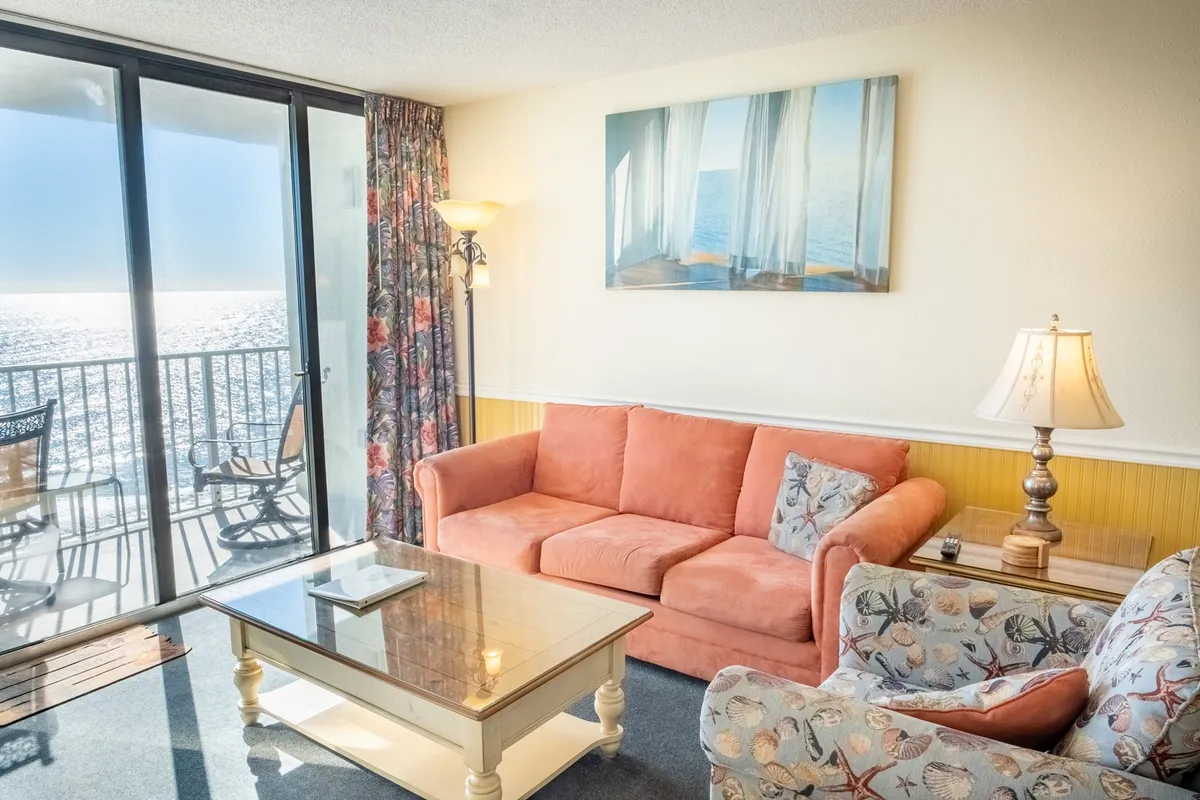 This image shows a bright, inviting living room with an orange sofa set against a backdrop of large glass doors that open up to the ocean. Artwork adds a touch of personality, while the view through the doors offers a direct connection to the private balcony and the waves beyond.