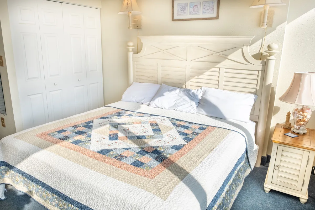 An inviting bedroom with a cozy bed topped with a patterned quilt, a nightstand and lamp, and framed pictures on the wall. Sunlight gently fills the space, enhancing its warmth and comfort.