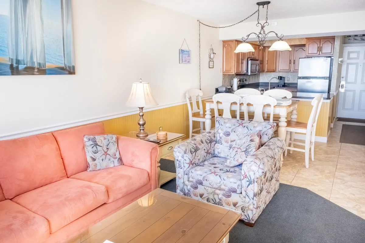 A cozy scene with a pink sofa, floral armchair, and a visible kitchen area in the background highlights the inviting atmosphere of the home. The kitchen is clearly part of an open-plan living area, underscoring the sense of togetherness.