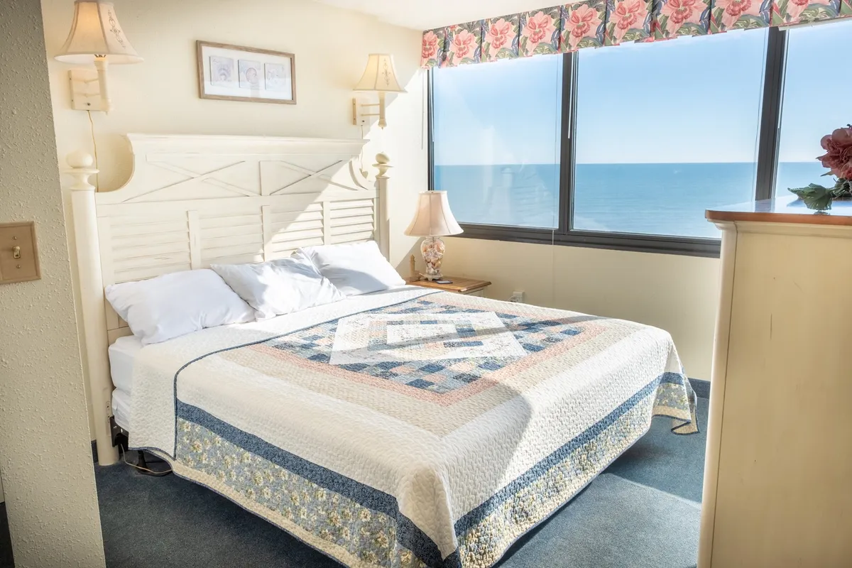 A serene bedroom featuring a comfortable bed with a quilt, large windows offering an ocean view, and a nightstand with a lamp. The room is filled with natural light and decorated in tranquil tones, creating a peaceful ambiance.
