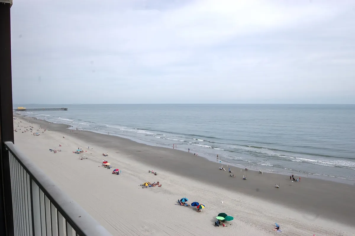 The image captures a sweeping view of the shoreline from the condo, with the sparkling Atlantic stretching to the horizon. Families and friends are visible on the sandy beach, relaxing under colorful umbrellas, sunbathing, strolling by the surf, and children playing or building sandcastles. The ocean is alive with gentle waves, and the scene radiates a laid-back, joyful beach vibe.
