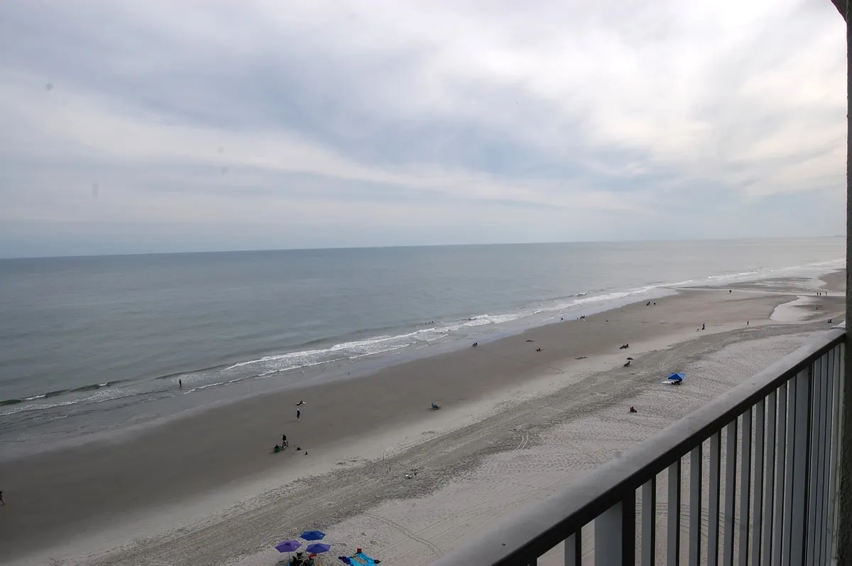 A wide, inviting view of Myrtle Beach's sandy shoreline with gentle ocean waves and people walking, sunbathing, swimming, and building sandcastles. Colorful umbrellas dot the scene, and the atmosphere is relaxed and lively, perfect for family activities.