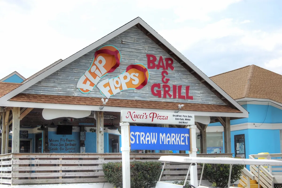An exterior scene highlighting the local flavor with visible signage for Flip Flops Bar & Grill, Nucci’s Pizza, and the Straw Market. The casual setting suggests opportunities for socializing, dining al fresco, and sampling local specialties—essential elements of a long-term stay experience.