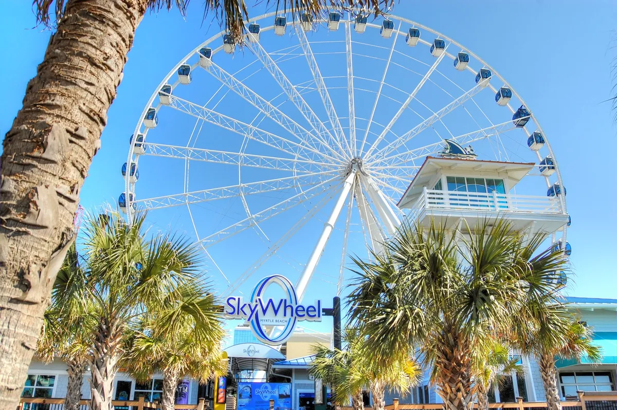 A dynamic exterior shot featuring the iconic Myrtle Beach SkyWheel rising above palm trees and vibrant entrance signage. The mood is joyful and bustling, with families and groups gathered near the entrance, ready for a memorable ride.