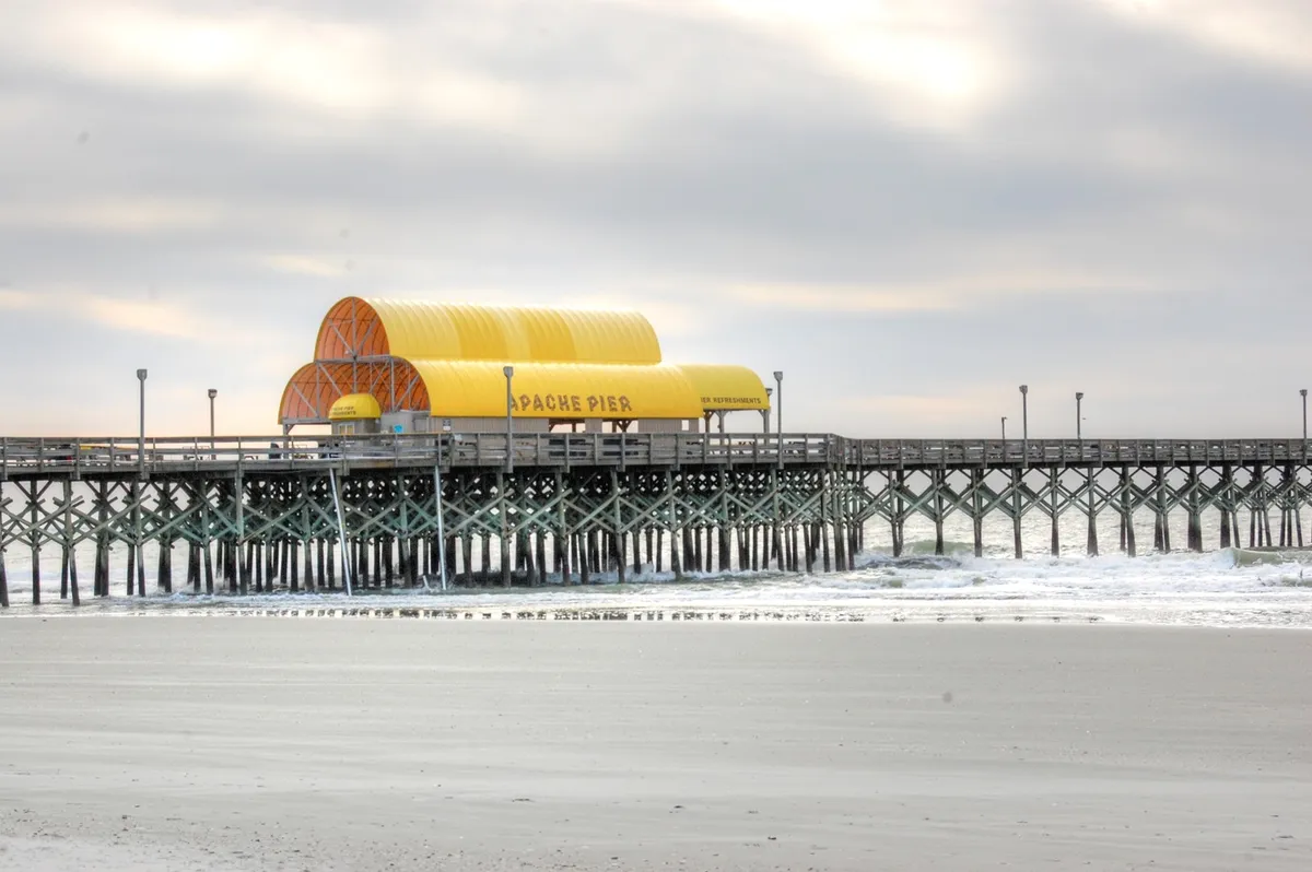 A bright, inviting exterior view of the iconic Apache Pier with its yellow roof, wooden structure stretching out to sea, and gentle ocean waves lapping the shore. The scene features people enjoying fishing, walking, and relaxing by the water, capturing the heart of Myrtle Beach's beautiful coastal setting.