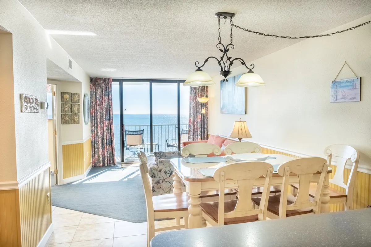 A cheerful dining area is pictured, complete with a dining table overlooking an expansive ocean view. Colorful curtains frame the window, and elegant light fixtures add charm, creating an inviting space for shared meals or morning coffee by the sea.