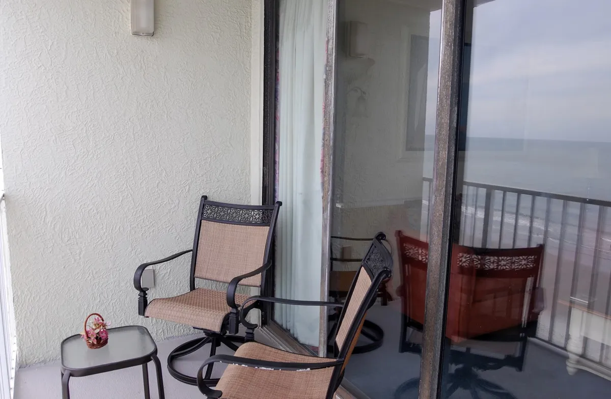 A private oceanfront balcony features two chairs and a small table, all facing an expansive view of the tranquil sea. Gentle waves can be seen in the distance, and the setup evokes a sense of intimacy and relaxation perfect for couples enjoying morning coffee or evening drinks together.